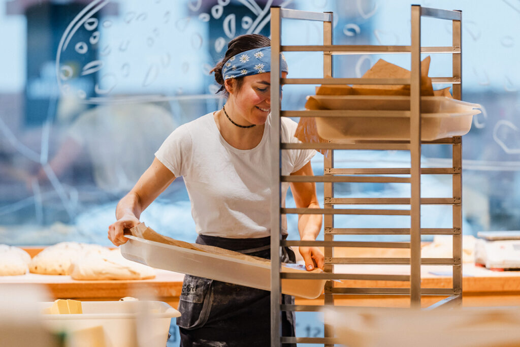 Bäckerin bei Till und Brot legt Teiglinge auf ein Blech – Blick in die gläserne Backstube einer modernen Handwerksbäckerei