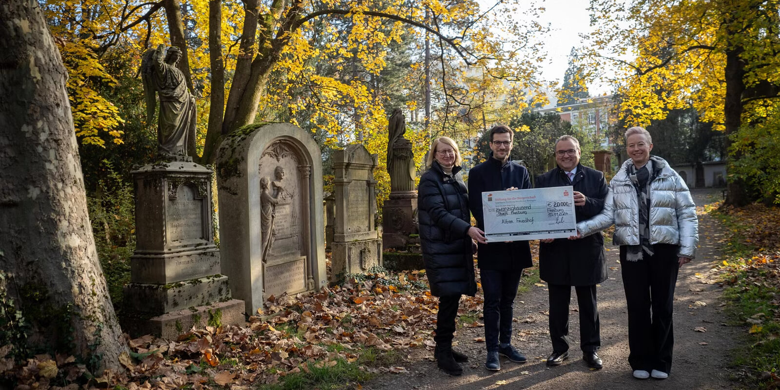 Andrea Katzer, Martin Horn und Daniel Zeiler übergeben einen symbolischen Spendenscheck über 20.000 Euro auf dem Alten Friedhof in Freiburg, umgeben von historischen Grabmälern und herbstlichem Laub