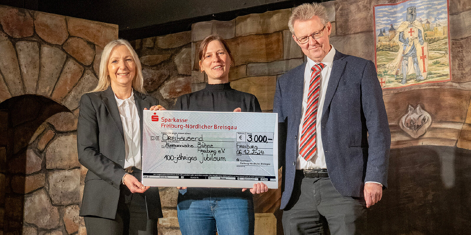 Beate Schwarz überreicht auf der Bühne einen symbolischen Spendenscheck über 3.000 Euro an Sandra Jettkandt von der Alemannischen Bühne und Bürgermeister Ulrich von Kirchbach anlässlich des 100-jährigen Jubiläums des Theaters