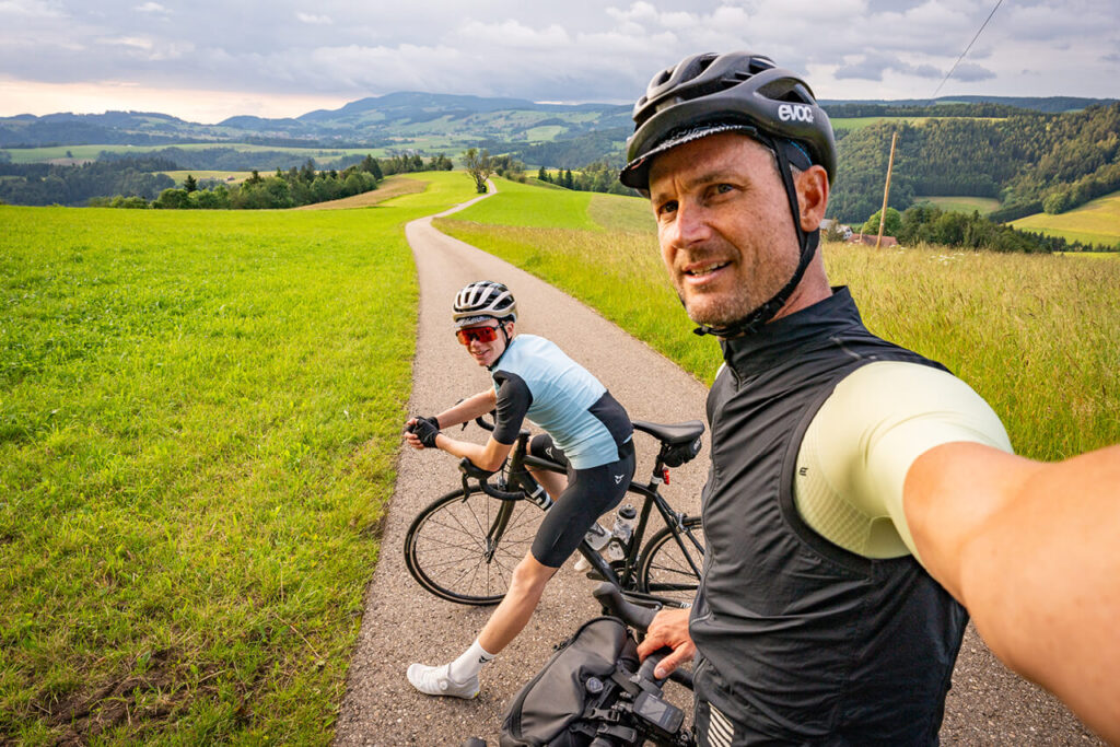 Baschi Bender und Sohn mit ihren Rädern auf einem asphaltierten Weg durch hügelige Landschaft, Baschi macht ein Selfie mit seinem Sohn im Hintergrund.