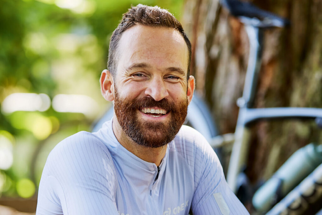Simon Geschke lächelt in die Kamera, im Hintergrund sein Fahrrad vor einem Baum.