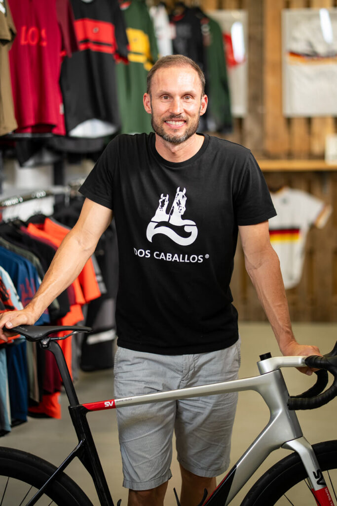 Marc Hanisch in seinem Freiburger Store für nachhaltige Radmode der Marke „Dos Caballos“, neben einem Rennrad stehend.