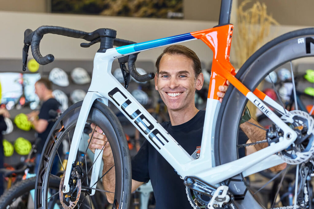 Jan Hättich von HOT.BIKE hält ein CUBE-Rennrad im Verkaufsraum mit Helmen und Fahrradzubehör im Hintergrund.