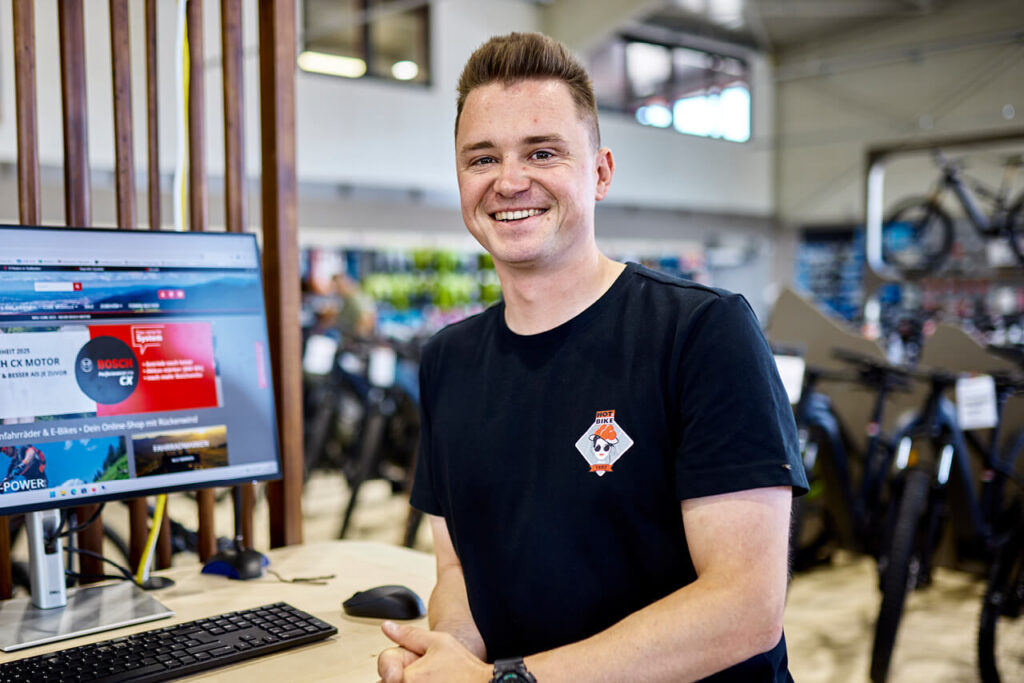 Sebastian Meyer von HOT.BIKE steht im Verkaufsraum vor einem Computer – im Hintergrund zahlreiche Fahrräder und Zubehör.