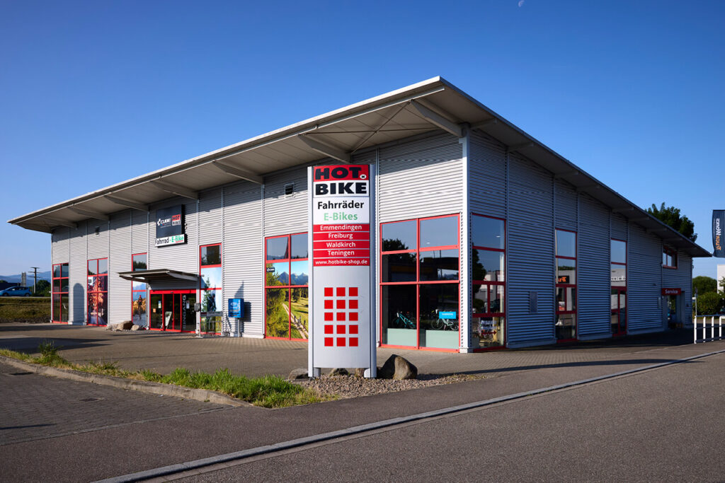 Außenansicht der HOT.BIKE-Filiale in Teningen mit großem Eingangsschild und roter Fensterrahmung bei Sonnenschein.