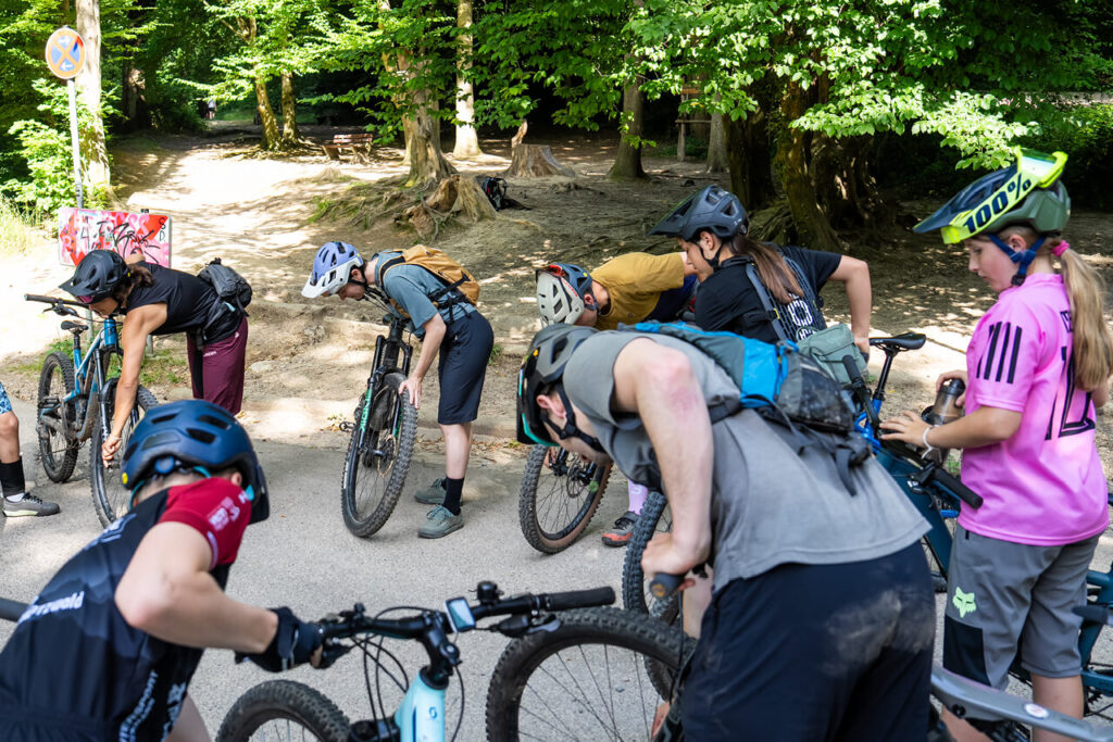 Gruppe junger Mountainbiker:innen beim Sicherheitscheck – sie kontrollieren gemeinsam ihre Räder vor dem Start des Jugendtrainings im Wald.