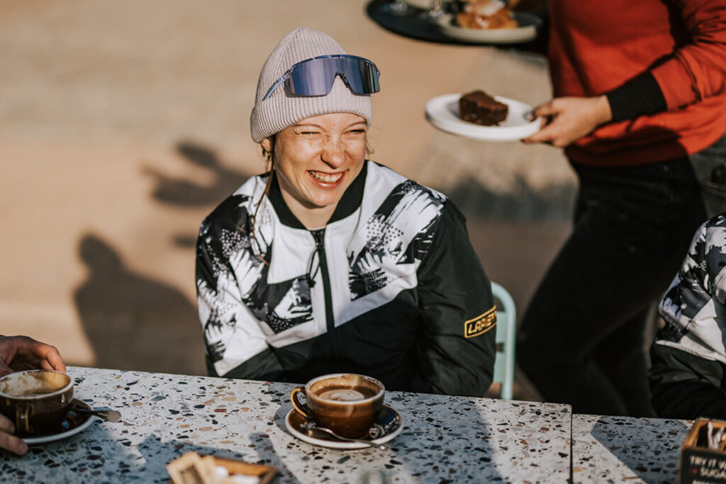 Junge Frau, Nina Graf, mit Radbrille und Mütze trinkt Cappuccino in der Sonne vor einem Café in Freiburg, trägt Sportjacke und lacht herzlich.
