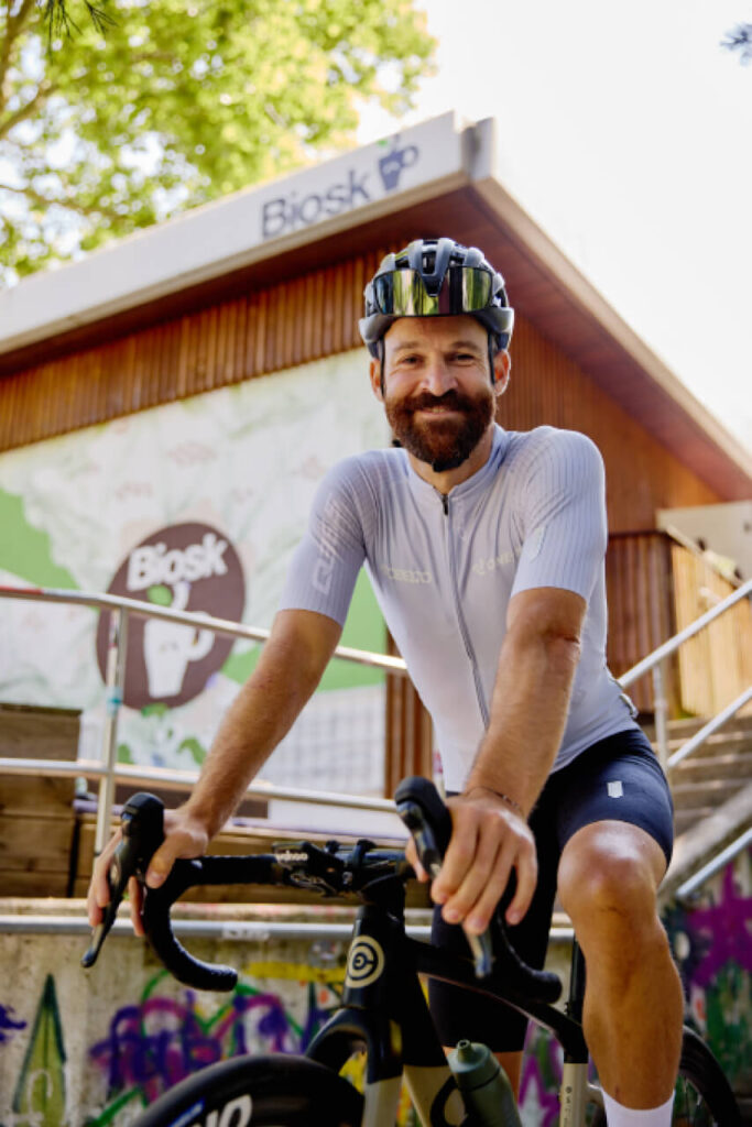 Simon Geschke sitzt in Radkleidung auf seinem Rennrad vor dem Biosk im Freiburger Osten und blickt lächelnd in die Kamera.