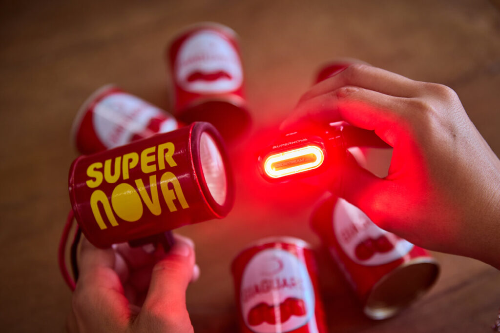 Zwei Hände halten Prototyp und aktuelle Fahrradlamp von Supernova Design, umgeben von Tomatenmarkdosen – Hommage an den Ursprung der Marke.