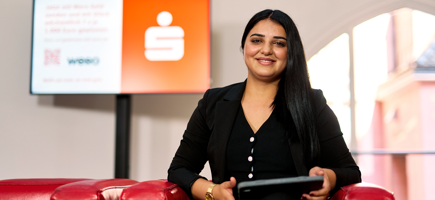 Auszubildende Shadiya der Sparkasse Freiburg sitzt im FinanzZentrum auf einem roten Sofa und hält ein Tablet, im Hintergrund ein Monitor.