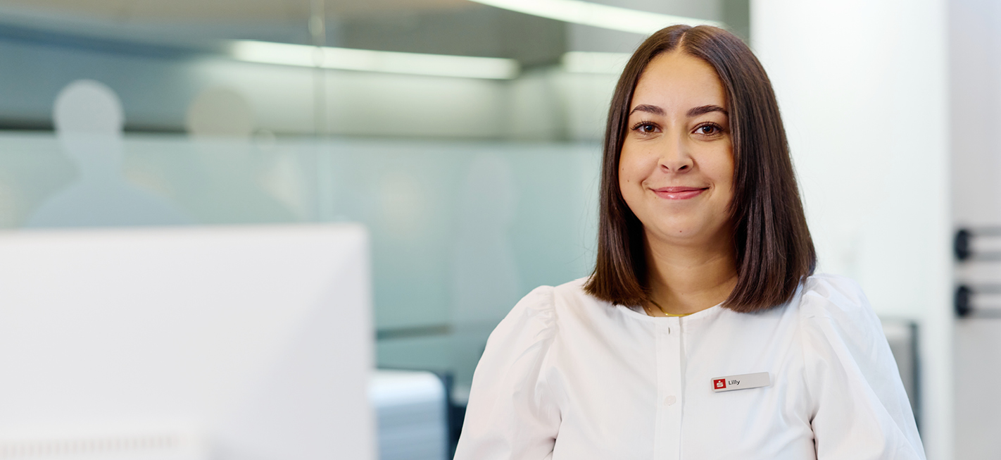 Auszubildende Lilly der Sparkasse Freiburg am Empfangsschalter, freundlich lächelnd, mit Namensschild und weißer Bluse.