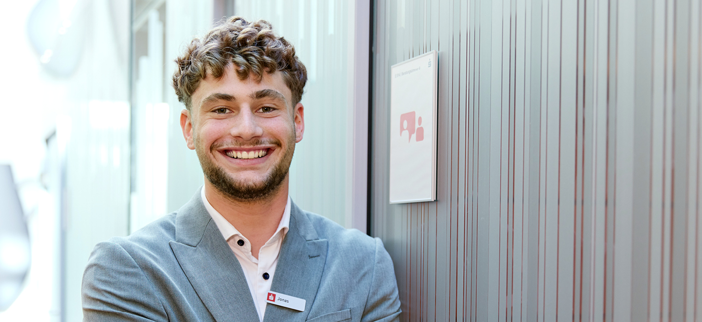 Auszubildender Jonas der Sparkasse Freiburg lächelnd im Beratungstrakt, mit Namensschild und grauem Sakko.