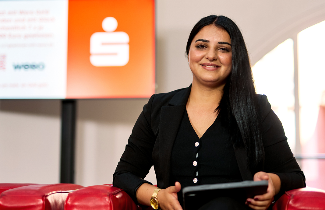 Auszubildende Shadiya der Sparkasse Freiburg sitzt im FinanzZentrum auf einem roten Sofa und hält ein Tablet, im Hintergrund ein Monitor.