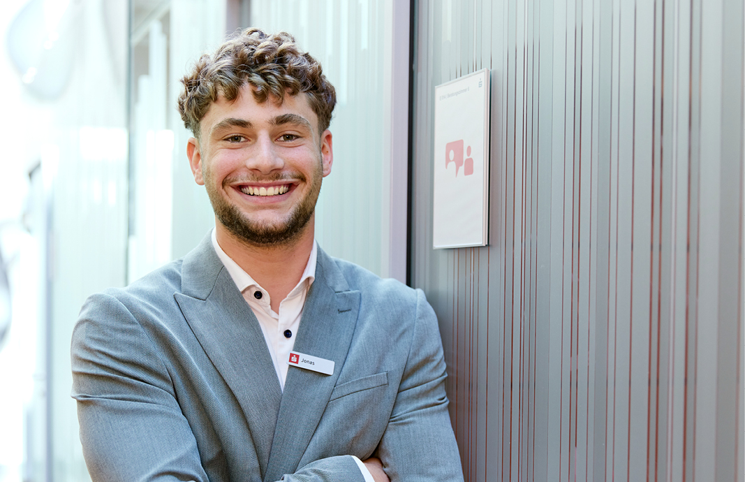 Auszubildender Jonas der Sparkasse Freiburg lächelnd im Beratungstrakt, mit Namensschild und grauem Sakko.