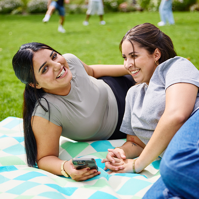 Die Auszubildenden Shadiya und Lilly liegen entspannt auf einer Picknickdecke im Grünen – beide lachen.
