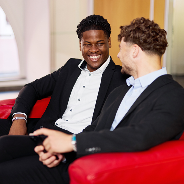 Die Auszubildenden Joys und Jonas der Sparkasse Freiburg im Gespräch auf einer roten Couch im FinanzZentrum.