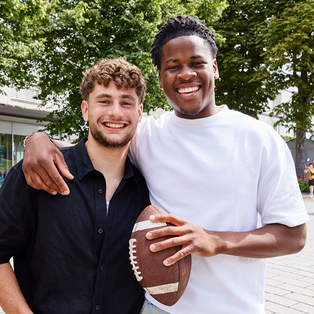 Die Auszubildenden Joys und Jonas der Sparkasse Freiburg posieren lachend mit einem Football im Freien.