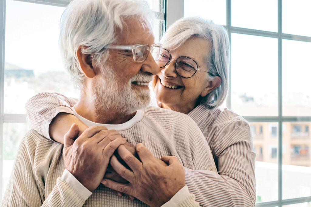 Älteres Paar steht lächelnd vor einem Fenster, sie umarmt ihn liebevoll von hinten, beide wirken glücklich und entspannt.