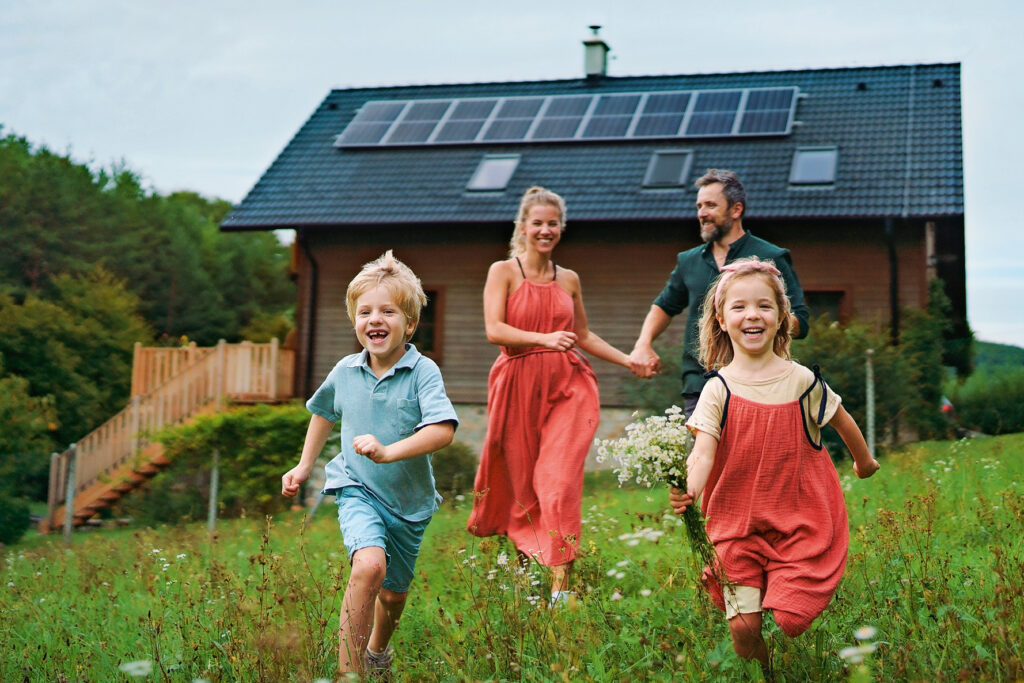 Familie mit zwei Kindern läuft lachend über eine Wiese vor einem Haus mit Solaranlage auf dem Dach.