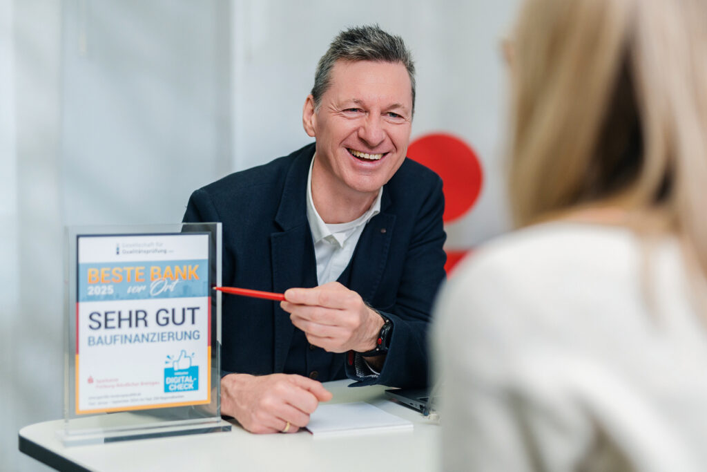 Bankberater im Gespräch mit Kundin, lächelnd, auf dem Tisch steht eine Auszeichnung als ‚Beste Bank vor Ort 2025‘.