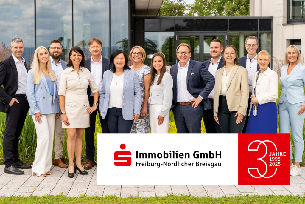 Team der S-Immobilien GmbH Freiburg-Nördlicher Breisgau steht vor dem Bürogebäude und blickt lächelnd in die Kamera. Jubiläumslogo 30 Jahre.
