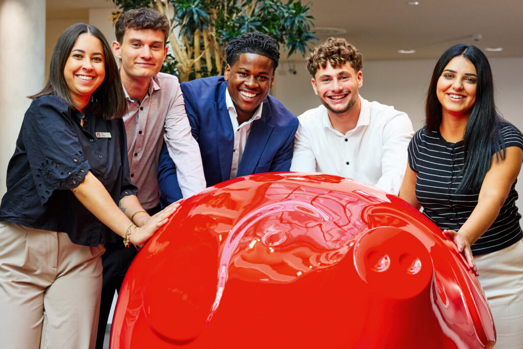 Fünf Auszubildende der Sparkasse Freiburg im FinanzZentrum lehnen lachend an einem übergroßen roten Sparschwein.