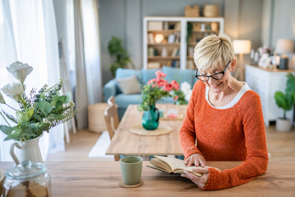 Frau mit Brille und orangem Pullover liest ein Buch am Holztresen in einem hellen Wohn-/Esszimmer