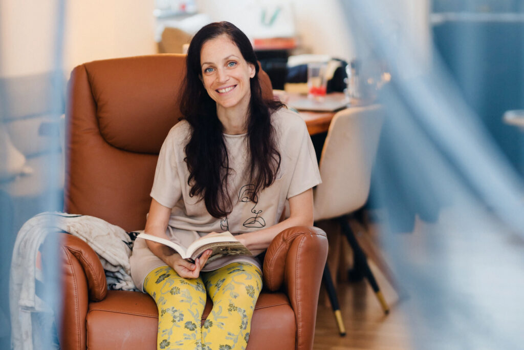 Frau sitzt in einem braunen Sessel, hält ein Buch in der Hand und lächelt in die Kamera