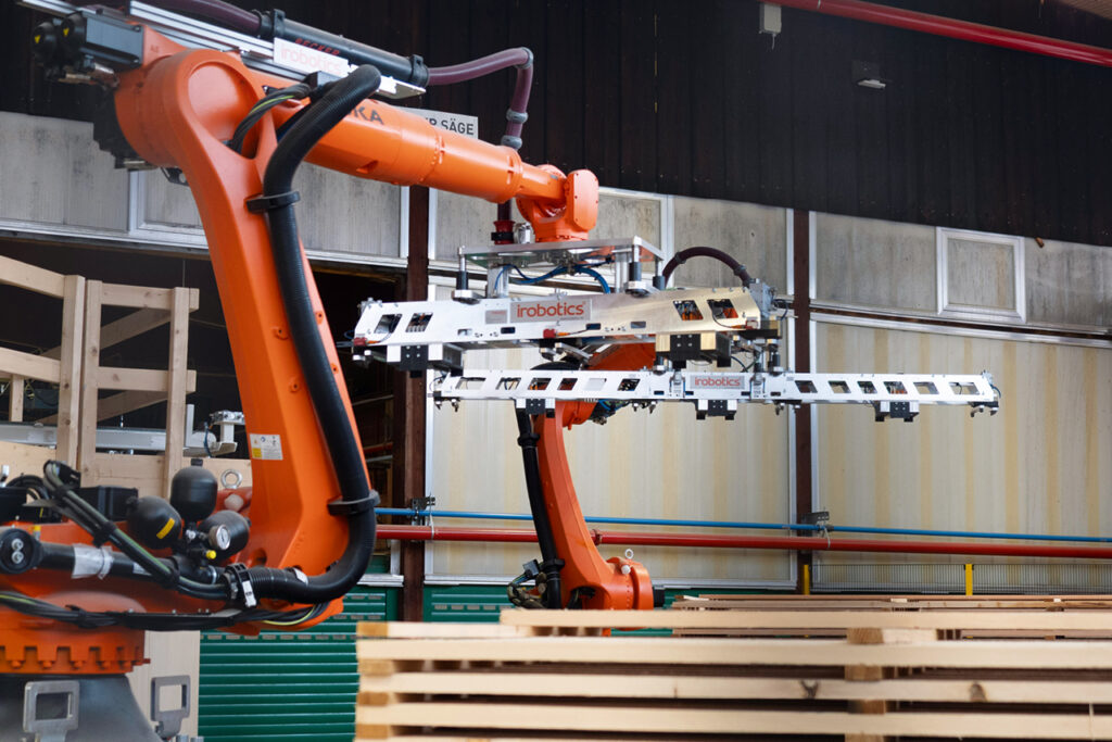 Ein orangefarbener Roboterarm bewegt in einer Produktionshalle Holzplatten mit einer Greifvorrichtung.