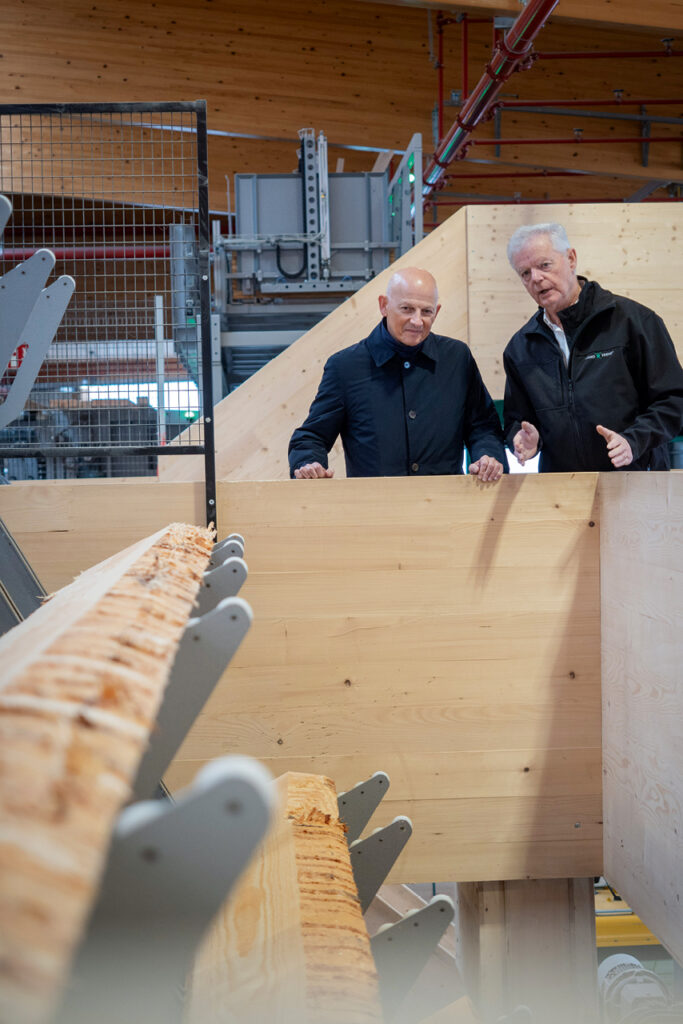 Zwei Männer stehen in einer Produktionshalle neben großen Holzelementen und sprechen miteinander.