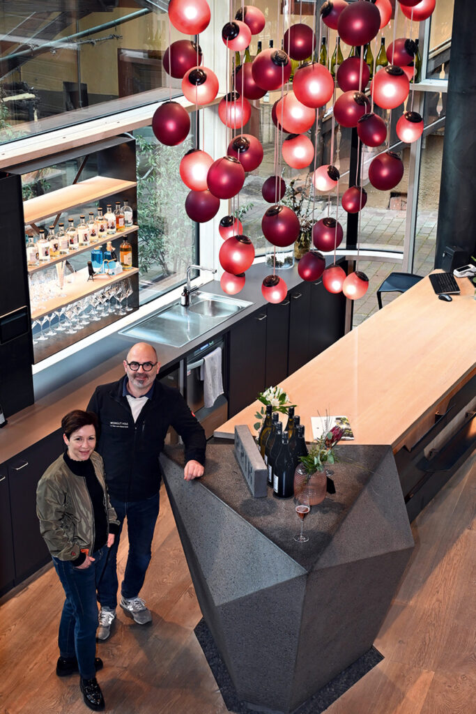 Andreas und Alexandra Hiss stehen im Verkaufsraum des Weinhauses Hiss unter einer Installation aus roten Leuchten neben der Theke.